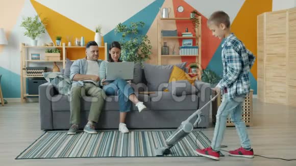父母在家里沙发上使用笔记本电脑时帮助儿子吸尘地板的缓慢运动视频的预览图