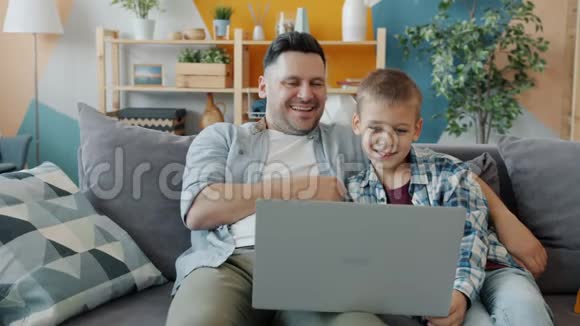 爸爸和儿子用笔记本电脑打字微笑着看着坐在沙发上的屏幕视频的预览图