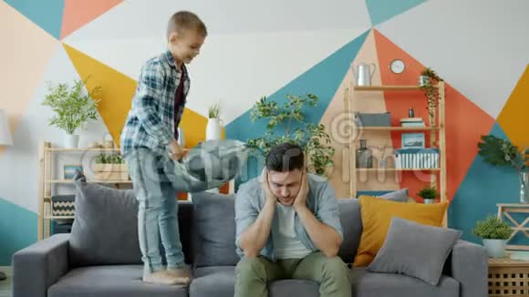 一个调皮的孩子在沙发上跳着枕头打他时爸爸头痛得很视频的预览图