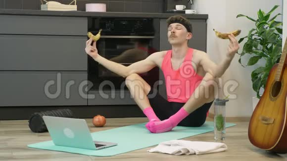 坐在莲花里的男人摆姿势手里拿着香蕉在厨房的家中健身垫上带胡子和粉色衣服的有趣的年轻人视频的预览图