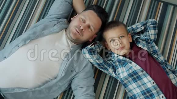 成年男子和可爱男孩躺在房子地板上一起聊天放松的肖像视频的预览图