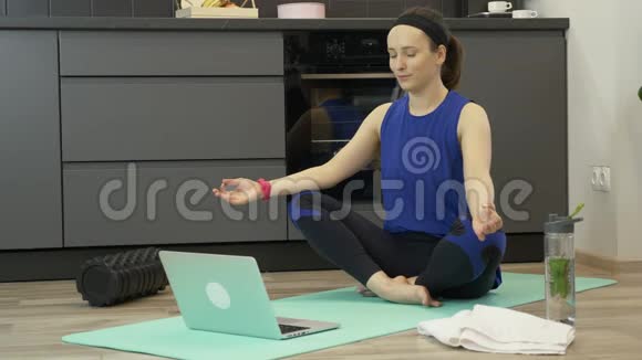 年轻女孩坐在莲花姿势在家里笔记本电脑上进行在线瑜伽课程女性在家中厨房练习瑜伽体式视频的预览图