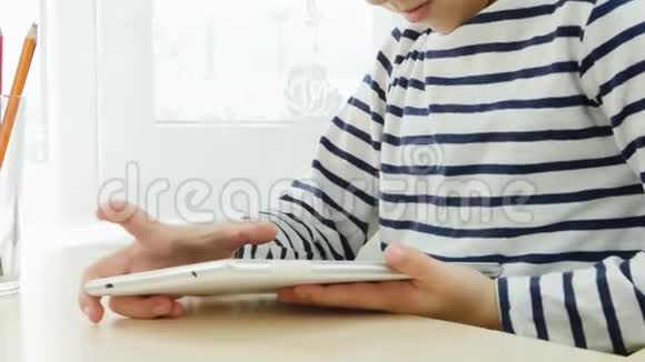 一个男孩坐在靠近窗户的桌子旁在社交网络中的平板电脑上玩耍孩子笑了快关门视频的预览图
