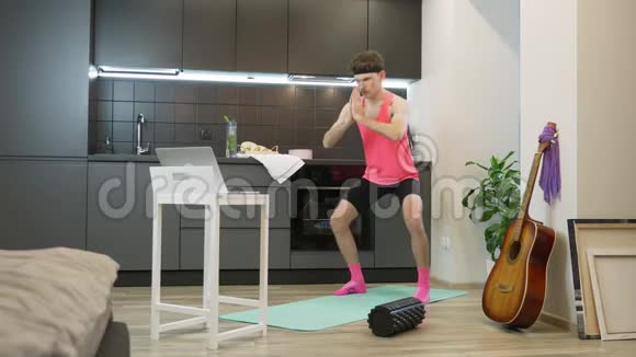 年轻人在家里用笔记本上的在线应用程序练习空手道有胡子和粉色上衣的有表现力的男人做锻炼视频的预览图