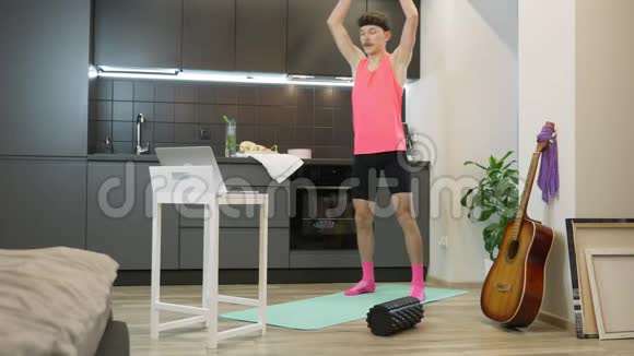 年轻的健身者在家里用笔记本电脑上的在线应用程序做运动穿粉色上衣的滑稽可笑的家伙视频的预览图