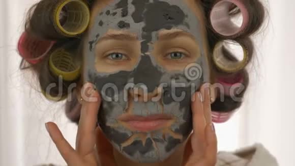 女人脸上戴着黑色粘土面具看着相机闭上眼睛头上有卷发的女性有美容程序视频的预览图