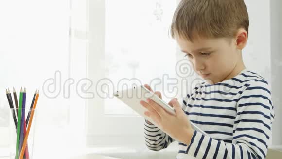 一个男孩坐在靠近窗户的桌子旁在社交网络互联网上的平板电脑上玩耍孩子正在看一个视频的预览图