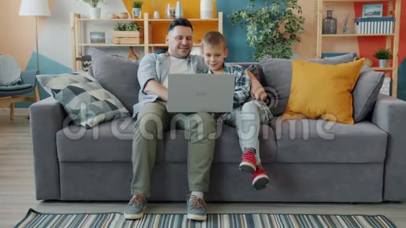 小男孩用电脑和父亲坐在工作室公寓的沙发上视频的预览图