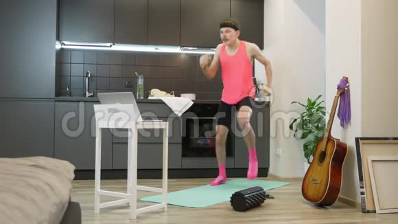 有趣的年轻人教练带着胡子在家里慢跑使用在线健身应用程序在笔记本电脑上穿粉色衣服的男性视频的预览图