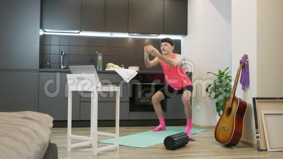 有趣的男子运动员在粉红色的顶部做蹲在笔记本上看在线健身课程在家里有趣的男子有氧运动视频的预览图