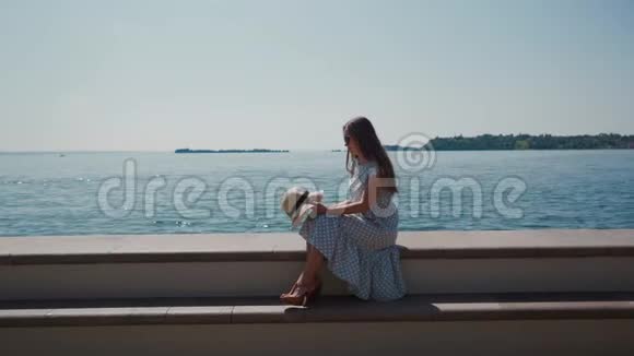 穿着浪漫礼服的女人坐在石墙上的蓝湖边手拿帽子视频的预览图