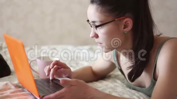 一个年轻迷人的女孩在家里躺在床上远程工作一个年轻可爱的女孩躺在床上用笔记本电脑工作视频的预览图