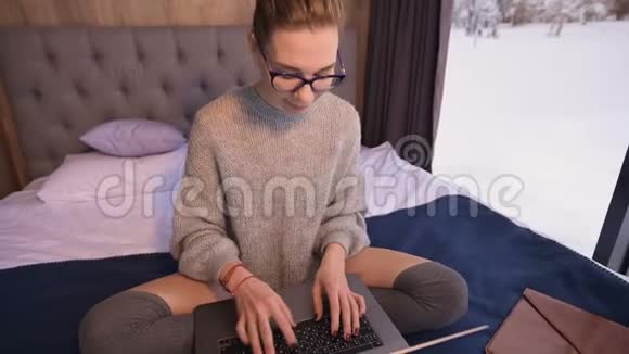 一个戴着眼镜的女自由职业者坐在乡间别墅的床上房子后面有一扇雪亮的窗户视频的预览图