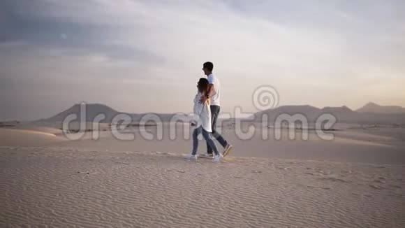 在阳光和蓝天的沙漠下拥抱的情侣在浪漫的关系中行走的慢镜头两个年轻人视频的预览图