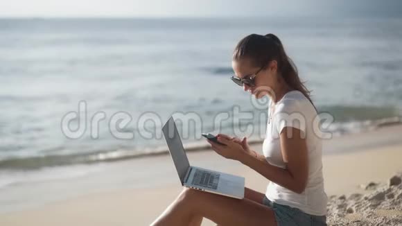 有笔记本电脑的女人坐在海边用手机进行视频通话视频的预览图