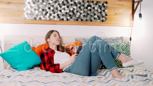 一个年轻迷人的白人女人躺在床上在现代卧室里微笑一边听着用现代耳塞沉思视频的预览图