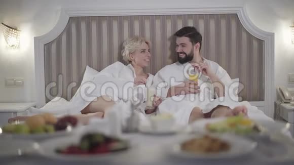 放松的高加索男人和女人躺在床上喝咖啡喝果汁说话入住酒店休息的快乐成年夫妇视频的预览图