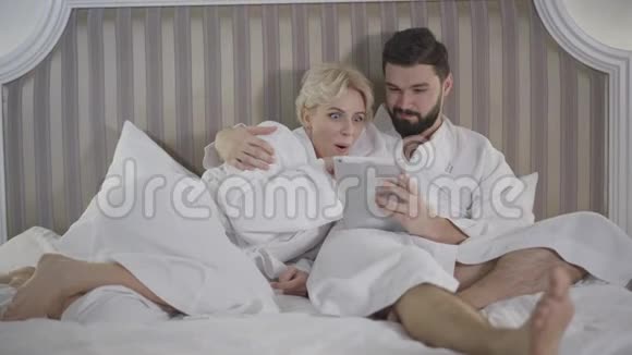 兴奋的高加索女人在平板电脑屏幕上向爱的男人展示一些东西一对美丽微笑的夫妇躺在床上视频的预览图