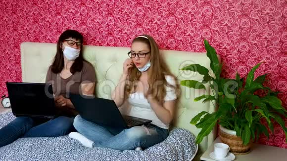 从家里远程工作两名戴着医用防护面罩的白种人妇女正在卧室的笔记本电脑上工作视频的预览图