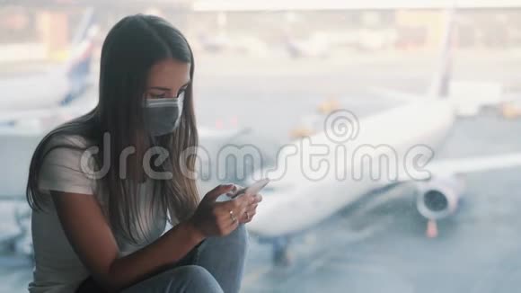戴防护面罩的女人坐在机场的窗户附近使用智能手机视频的预览图