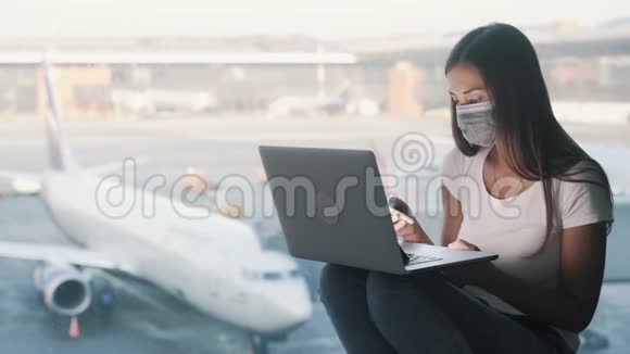 戴防护面罩的女孩在机场使用笔记本电脑和电话交谈视频的预览图