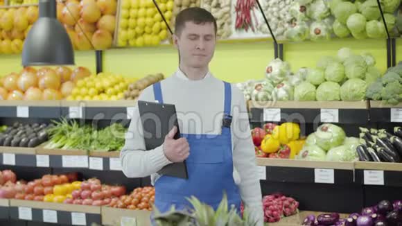 年轻积极的白种人拿着文件夹和茄子在杂货店摆姿势微笑的制服工人的画像视频的预览图