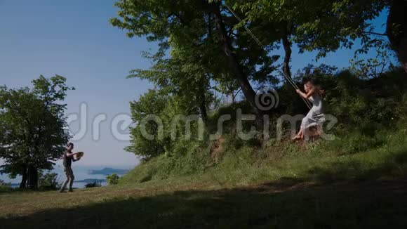 在一个自然公园里一个女孩在秋千上荡秋千一个男人把帽子扔在女孩的头上视频的预览图
