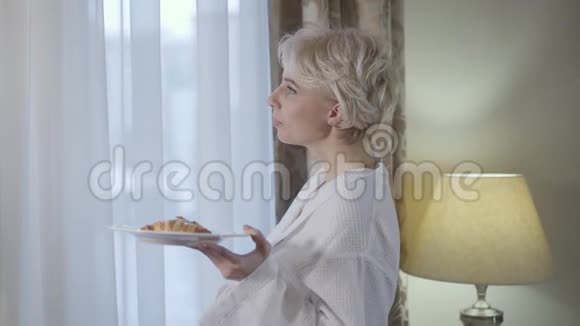 一边看着美丽的金发女人看着窗外吃着新鲜美味的牛角面包年轻的白种人女士视频的预览图