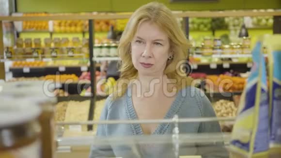 自信的成年白种人女人在超市的货架上张望的肖像集中的金发家庭主妇视频的预览图