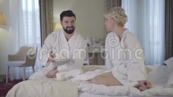 酒店房间里浴袍中快乐的成年男女碰杯年轻的白种人夫妻在高海拔地区休息视频的预览图