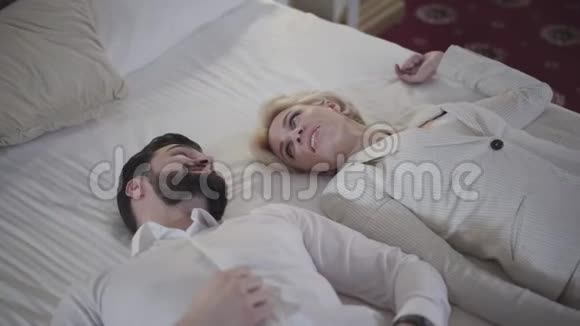 年轻快乐的白人夫妇穿着优雅的白色衣服躺在床上说话微笑的金发女人和长胡子的男人视频的预览图