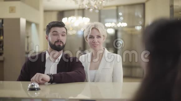 充满自信的白种人夫妇在酒店与接待员交谈的肖像年轻的黑发男子和金发女子视频的预览图