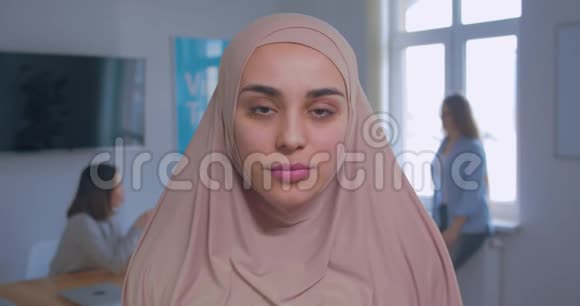 一位年轻漂亮严肃的穆斯林女孩在头巾办公室背景下的特写肖像视频的预览图