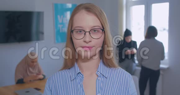 一位年轻美丽的白种人戴着眼镜的金发女郎在办公室背景上的画像视频的预览图
