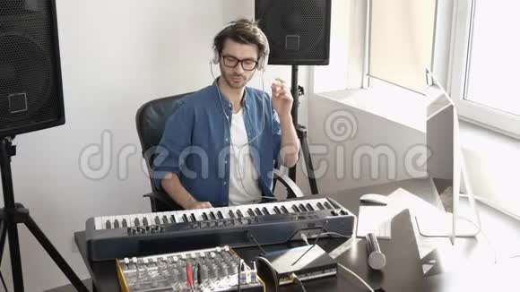 录音室的年轻人坐在桌子上使用混合控制台通过耳机听音乐视频的预览图
