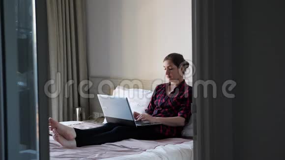 女士坐在白色的床上在灰色的现代笔记本电脑上打字视频的预览图