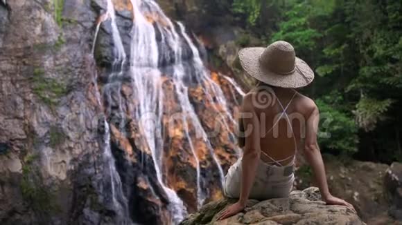 一个戴着草帽的漂亮女孩坐在岩石上背对着相机看着瀑布视频的预览图
