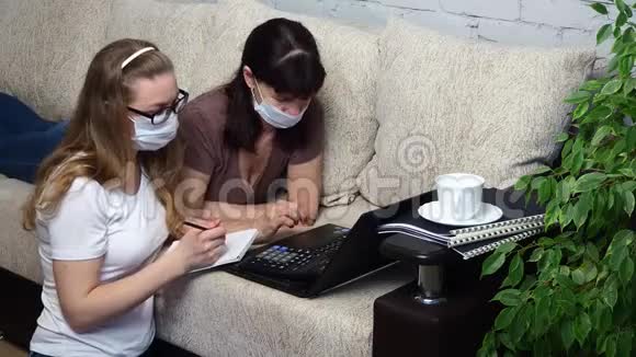 从家里远程工作两名戴防护口罩的妇女一起在客厅沙发上的笔记本电脑上工作视频的预览图