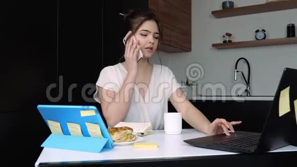 检疫期间厨房里的年轻女子在电话上聊天在笔记本电脑上工作在家远程工作表视频的预览图