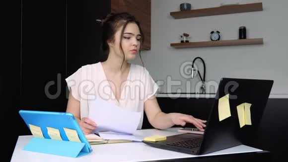 检疫期间厨房里的年轻女子带文件和笔记本电脑的远程工作打电话给智能手机谈谈视频的预览图