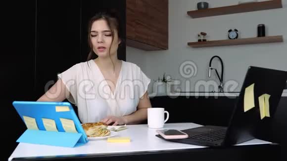 检疫期间厨房里的年轻女子使用平板电脑和笔记本电脑远程工作桌上有一杯饮料的三明治视频的预览图