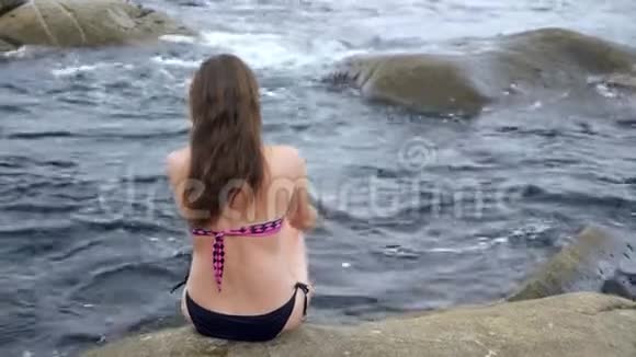 穿着时髦比基尼的女人坐在靠近深海背面的岩石上视频的预览图