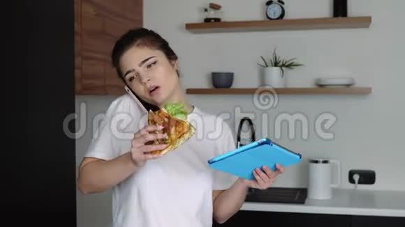 检疫期间厨房里的年轻女子女孩打电话吃三明治手拿平板电脑远程工作视频的预览图