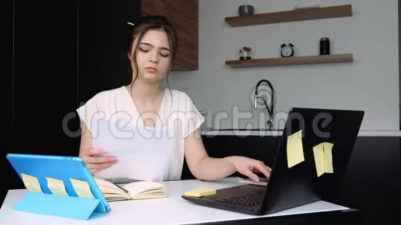 检疫期间厨房里的年轻女子严肃集中的女孩在笔记本电脑键盘上打字手拿文件视频的预览图