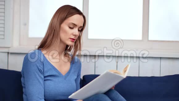 自信的年轻女人坐在靠近窗户的沙发上看书中型摄影机视频的预览图