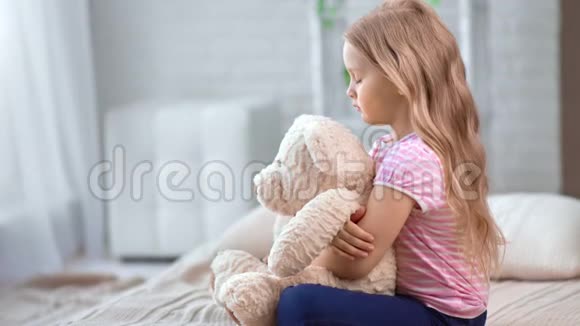 可爱的小女孩心烦意乱地抱着玩具泰迪坐在舒适的卧室床上4k龙红相机视频的预览图