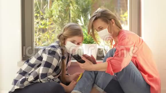 戴着医用口罩的妈妈和女儿正坐在窗边拿着手机隔离期间的消遣视频的预览图