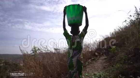 从一个遥远的乡村水处带着一只沉重的水桶回家的非洲少女的后景视频的预览图