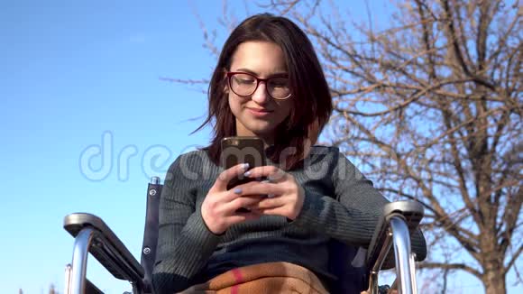 坐在轮椅上带着电话的年轻女子自然界中的残疾女孩在智能手机上通信视频的预览图