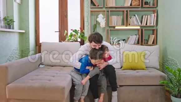 年轻的父亲和他的两个孩子笑着一起在客厅的沙发上玩耍他们享受着这一刻视频的预览图
